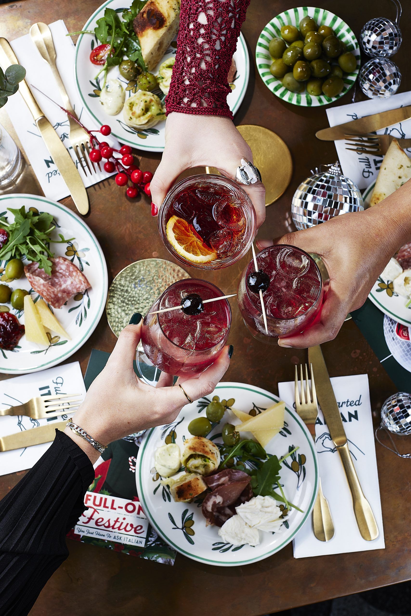 Three people cheersing their drinks over an abundant table. Each person has a plateful of Italian-style antipasti; salami, bread, salad and olives. 
