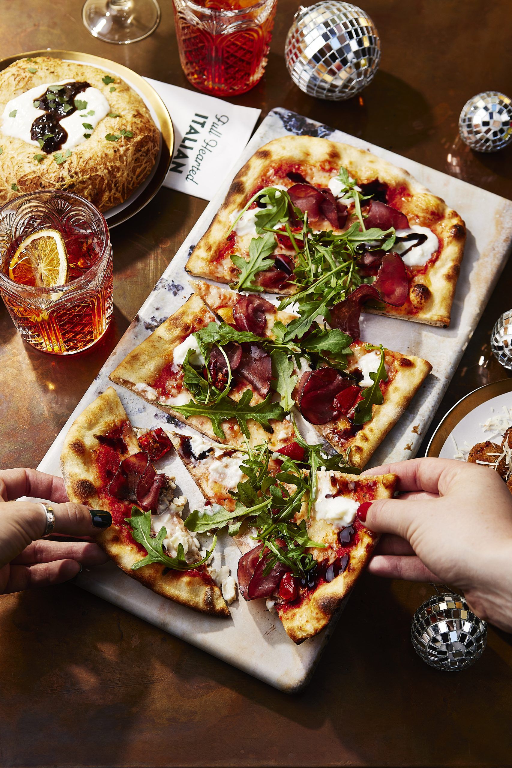 Two people grabbing a slice of pizza. The table decor has been adorned with Christmassy elements like baubles & mini disco balls. 
