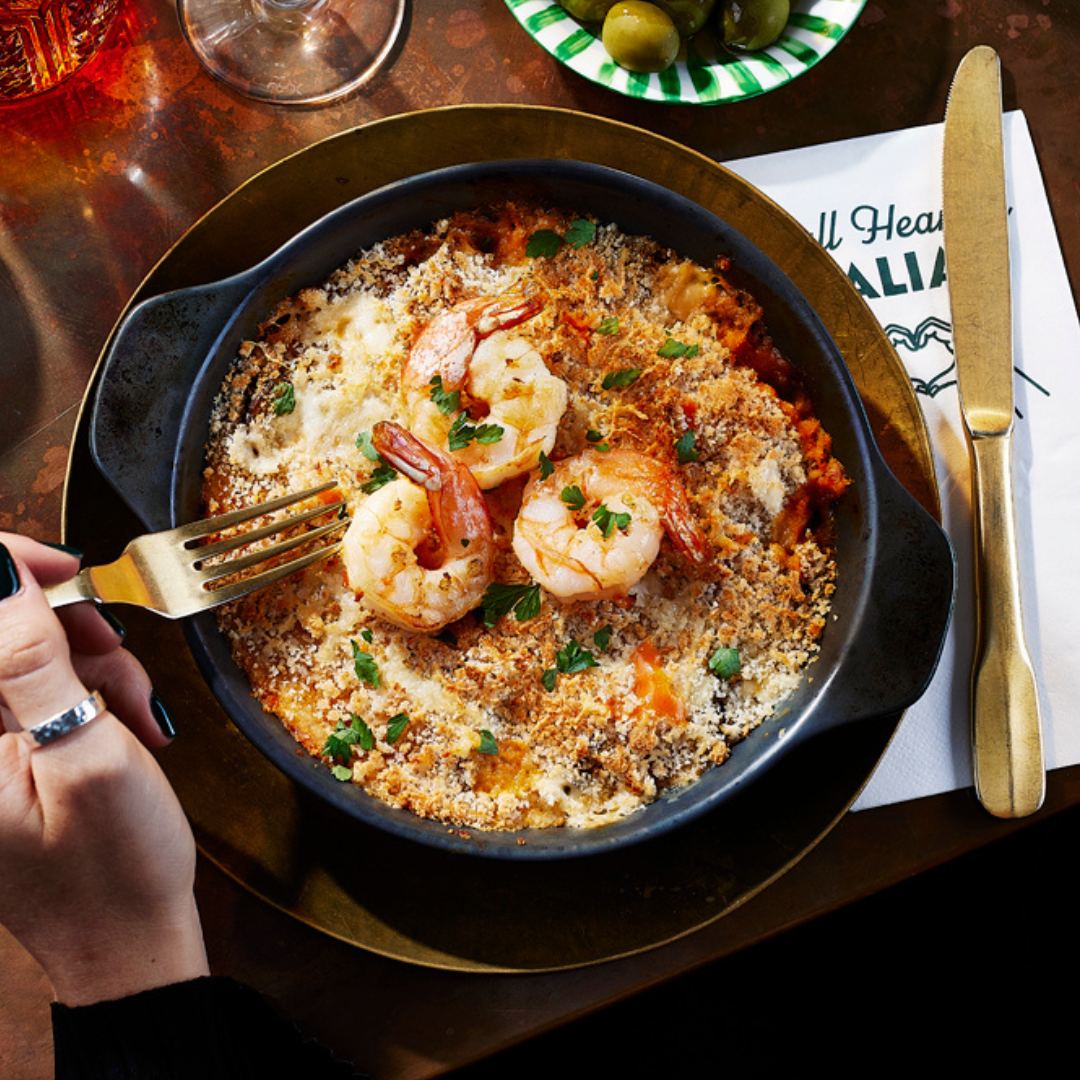 A photograph of a Seafood Gratin, oven baked with three King Prawns on top. The seafood gratin has been topped with crispy breadcrumbs and there's somebody digging in with a fork. 